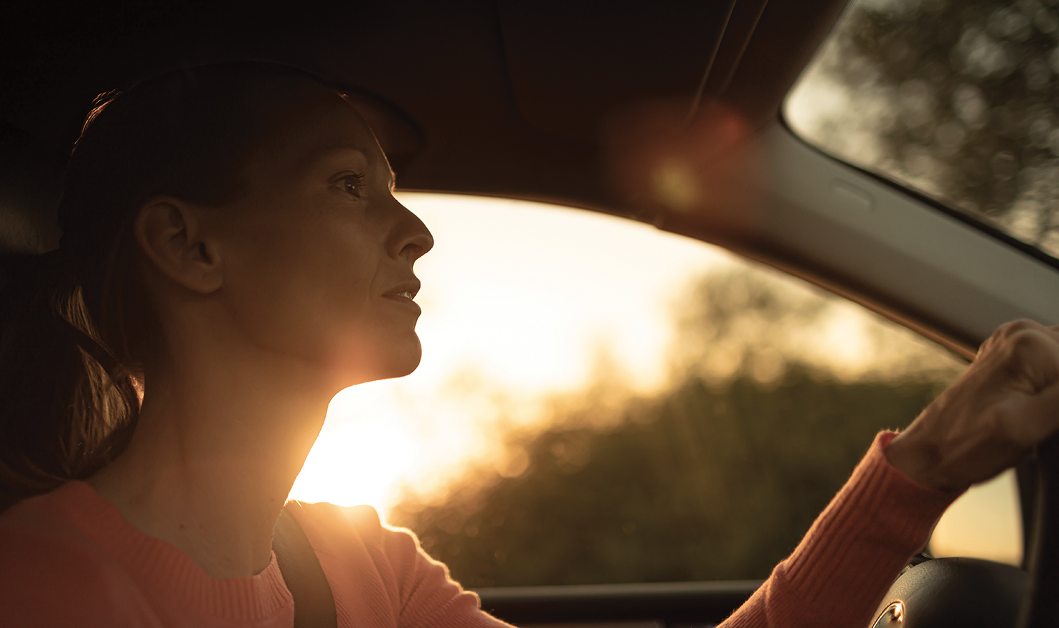 close up on woman's face , focused on driving a car, with sunset glowing behind her shoulder 