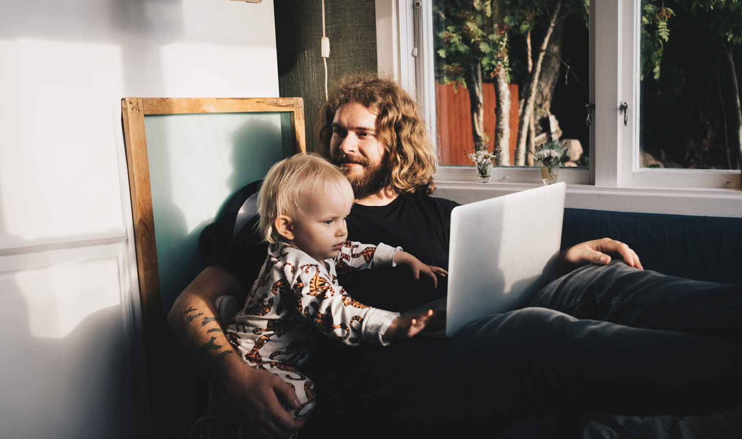 A father sitting on the sofa with his laptop on his lap, and his toddler acting curious